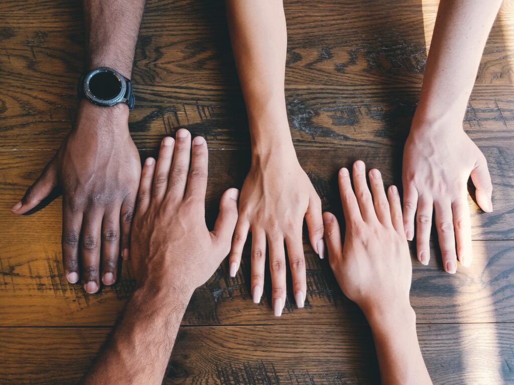 Five hands on a wood table of varying skin colors ranging from darker skin to lighter.