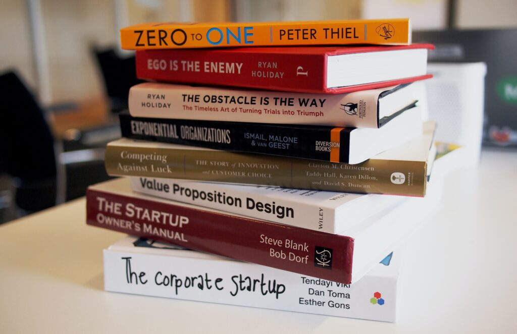 A stack of business books on a desk: Zero to One, Ego is the Enemy, Obstacle is the Way, Exponential Organizations, Competing Against Luck, Value Proposition Design, The Startup Owners Manual, The corporate startup.