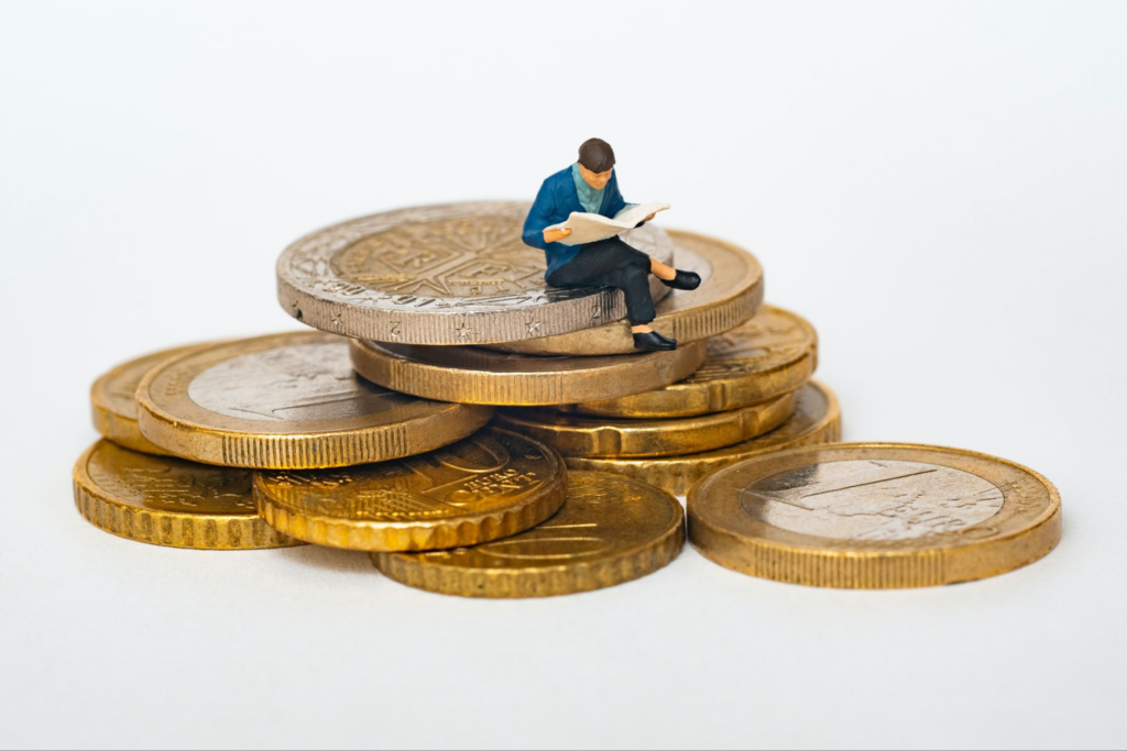 A tiny figurine of a man reading the newspaper sitting on a pile of coins.