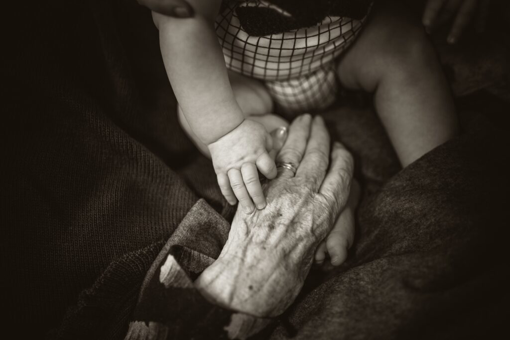 A black and white photo of a baby holding the hand of an elderly person.