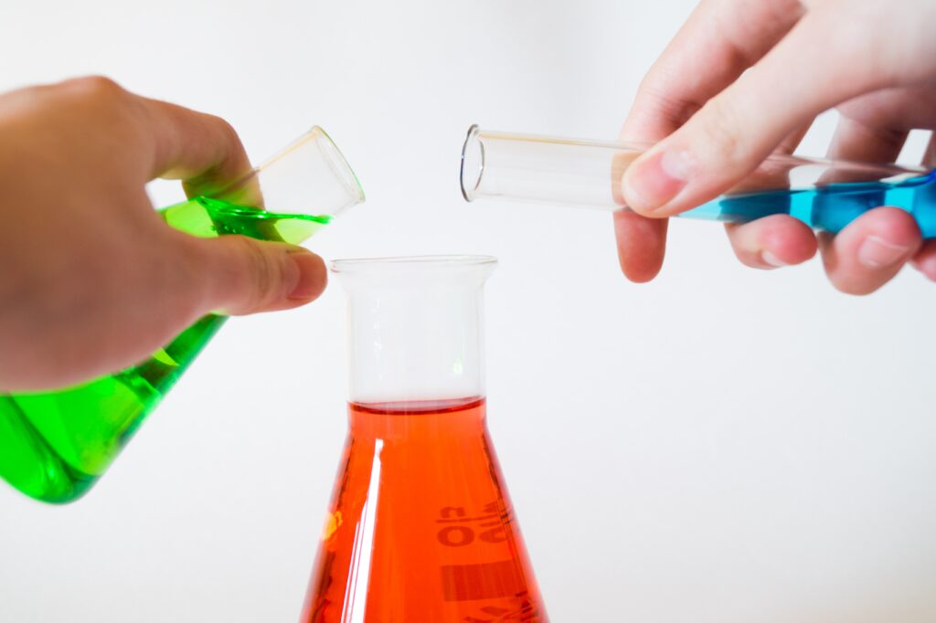 Hands pouring a blue test tube of liquid and a green flask of liquid into an (already far too full) flask of orange fluid.