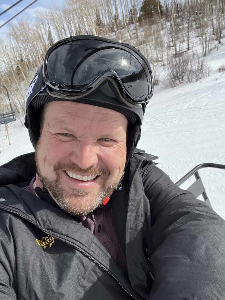 A middle-aged man trying something new - riding a ski lift up a mountain for the first time. He has the smile of someone who has no clue what he’s doing or what he’s gotten himself into.